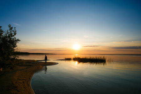 Sun shining on horizon over huge lake. Lonely person walking along shoreの写真素材