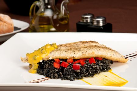 Plate of halibut fillet over a bed of rice with squid ink, capers tomato and mustard in a restaurant setting with copyspace.の写真素材
