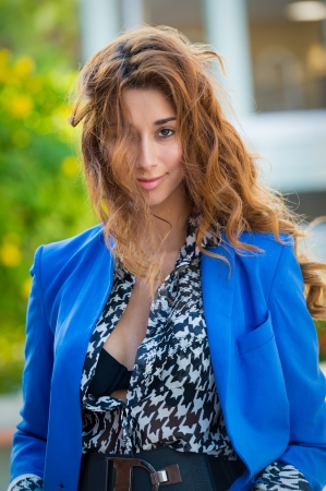 Beautiful elegant young woman dressed in trendy and stylish blouse standing outdoors looking at the camera smiling and long curly brunette hairの写真素材