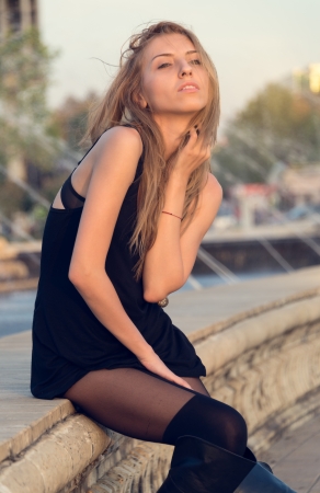 Sexy attractive woman in a short black dress and long boots posing sitting on a wall outdoors with her face raised to the evening sunlightの写真素材