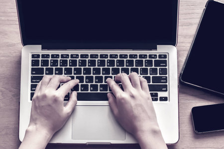 Close up of two hands typing on keyboard of a laptop next to a digital tablet and mobile phone devices on the table.の写真素材