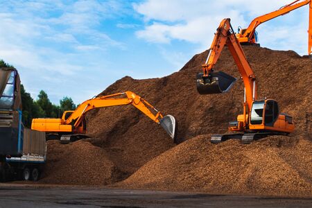 Pile of wood chips, backhoe scoping chips onto truck with blue sky backgroundの写真素材