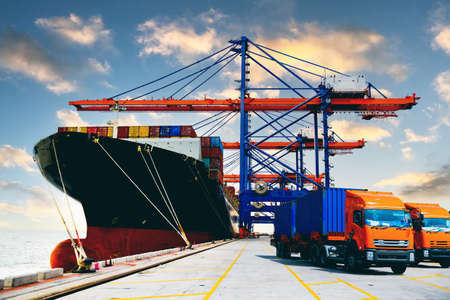 A fleet of heavy-duty trucks receives cargo containers from a huge ship under a quay crane. Container logistic import export and transport industry background.の写真素材