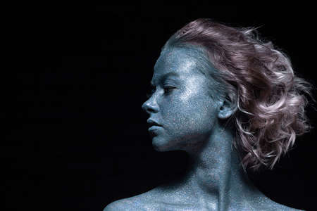 a girl with a blue face in profile in sparkles close-up against a black wall looks forwardの写真素材