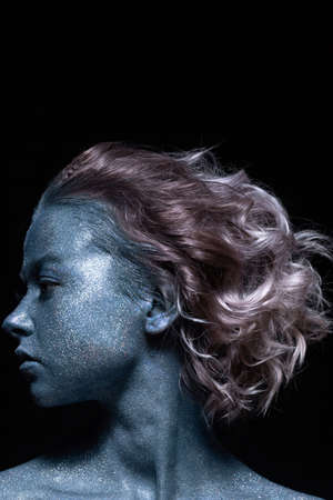 a girl with a blue face in profile in sparkles close-up against a black wall looks forward