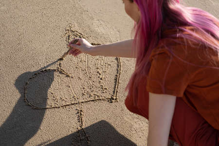 woman with pink hair draws a heart with a stick on the sandの写真素材