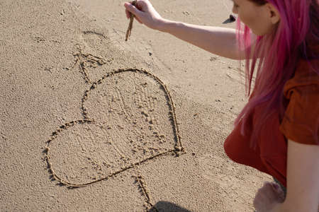 woman with pink hair draws a heart with a stick on the sandの写真素材