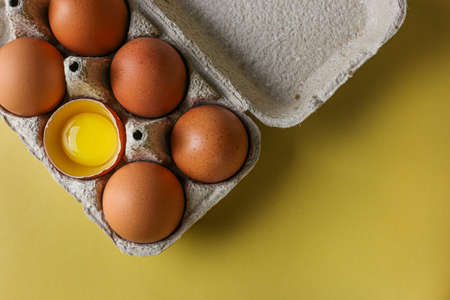 Fresh brown eggs and a broken egg with a yolk in a recycled paper eco tray on a light yellow...
