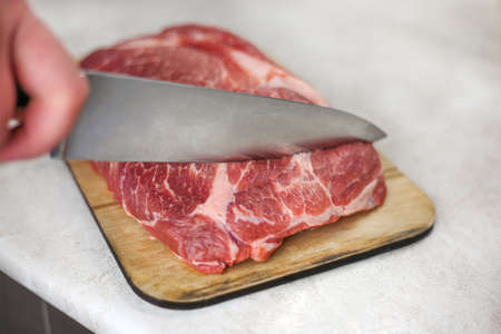 Cutting with a large knife a fresh piece of meat on a wooden cutting board on a light kitchen tableの写真素材