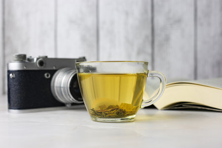 Brewed herbal green tea in a transparent cup next to an open book and a vintage camera on a light backgroundの写真素材
