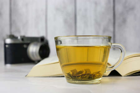 Brewed herbal green tea in a transparent cup next to an open book and a vintage camera on a light backgroundの写真素材