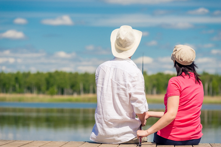 family couple with a fishing rod sitting on a wooden pier near the lake, rear viewの写真素材