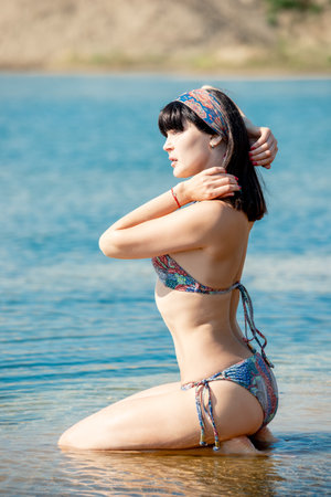 Beautiful slender brunette girl poses sitting in the water near the beach shoreの写真素材