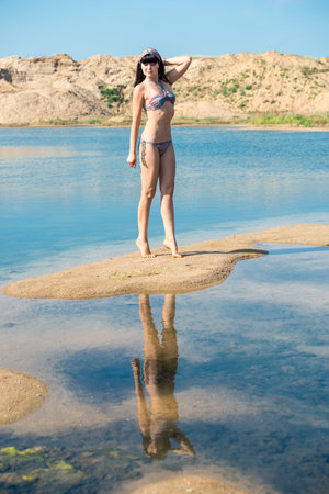 Beautiful slender brunette girl in a swimsuit poses on the sandy shore of a reservoir in sunny weatherの写真素材