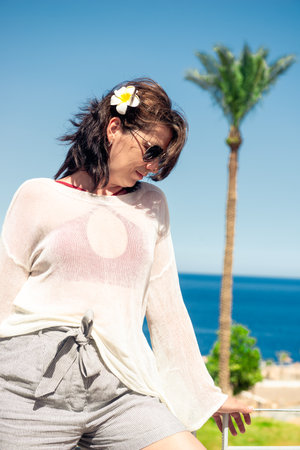 Beautiful girl in a white tunic poses against the backdrop of a warm tropical sea in sunny weatherの写真素材