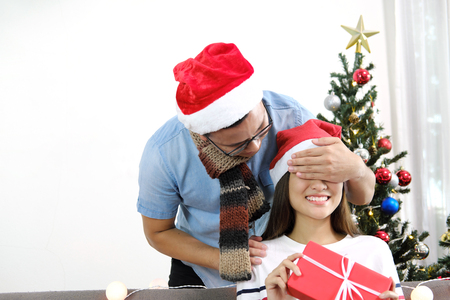 Young man in santa hat happy surprise closing girlfriend eyes with hands with gift box in Christmas holidays.の写真素材