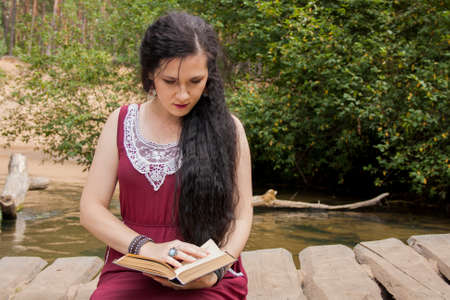 Girl sitting on a wooden bridge over the river and reading a bookの写真素材