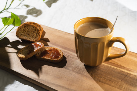 Light breakfast of toast with jam and a large orange cup of coffee with milk on a wooden board in white background.の写真素材