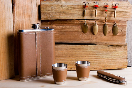 Hip stainless flask, cups and knife decorated leather on the table. Fishing metal baits on wooden background.の写真素材
