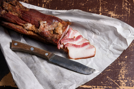Close up view slices of smoked bacon and knife on the white packaging paper. Naturmort in a rustic style on brown vintage background.の写真素材