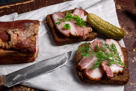 Close up view slices of smoked bacon on the piece rye bread with herbs, several pickles, old knife with wooden handle - rustic style on brown vintage background.の写真素材