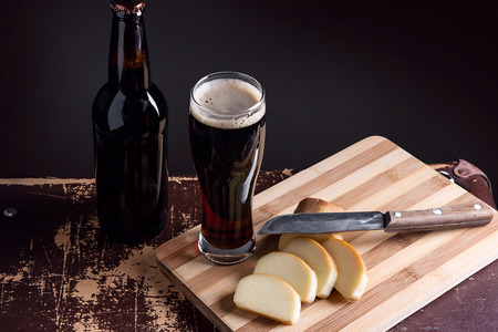 Glass with dark beer and full bottle of beer on dark background. Slices of smoked cheese with vintage knife on cutting board.の写真素材