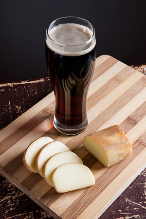 Full glass of dark beer with foam on vintage background. Slices of smoked cheese on cutting board.の写真素材