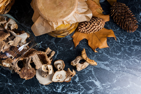 Two glass jars with wild mushrooms on black marble background. One jar with dry mushrooms and one with pickled mushrooms. Several dry white wild mushrooms on the table. Yellow wooden basket on back background, several dry oak leaves, acorns and fir cones.の写真素材