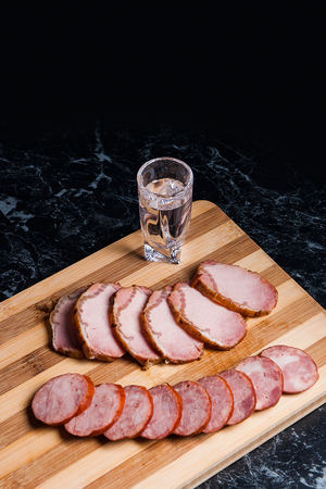 Shot glasses with vodka. Slices of smoked meat or ham and smoked sausage on wooden cutting board. Composition on black marble background.
の写真素材