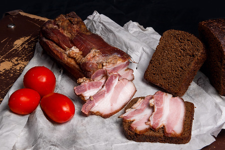 Close up view slices of smoked bacon with rye black bread on the white packaging paper. Slices of smoked bacon on the piece rye bread and red tomatoes - rustic style on brown vintage background.の写真素材