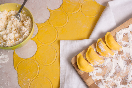 Close up view of several ready for boiling vareniki, dumplings, pierogi with cottage cheese or curd on wooden cutting board with flour. Fresh prepared yellow rolled out dough with white flour and green bowl with cottage cheese or curd, wooden rolling pin,の写真素材