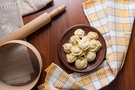 Boiled homemade meat dumplings served in clay plate with creamy sauce. Composition in rustic style on wooden background with boiled Ukrainian ravioli and rolling pin, sieve on brown cloth.の写真素材