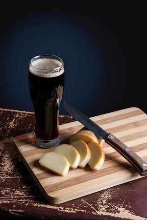 Full glass of dark beer with foam on vintage background. Slices of smoked cheese with vintage knife on cutting board.の写真素材