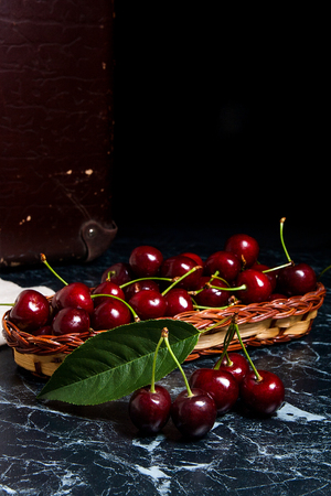Close up view of several red sweet cherries and big green leaf of cherry tree on the marble table and ripe sweet cherries in yellow wooden basket on background. Composition in rustic style - organic red sweet cherries in vintage wooden basket on dark marbの写真素材