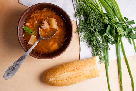Borsch - traditional Ukrainian and Russian red beetroot soup with red beets in clay bowl with metal spoon in it. Herbs - dill, green onion, parsley and white wheat bread around the plate. Ukrainian tomato soup borsch on wooden background.の写真素材