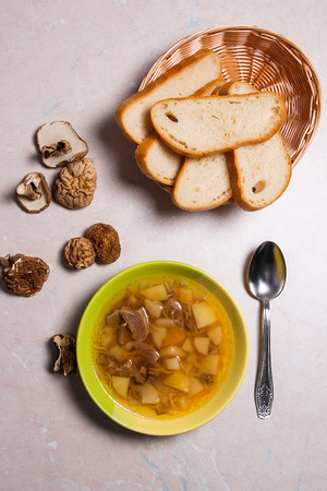 Mushroom soup in green plate with metal spoon on a light stone background. Several dried porcini or white  wild mushrooms on brown cloth. Slices of white bread in basket.の写真素材