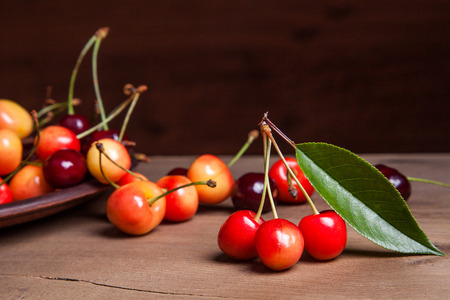 Clay plate with berries of yellow and red sweet cherries and several fruit with green leaf of cherry tree on vintage wooden backgroundの写真素材