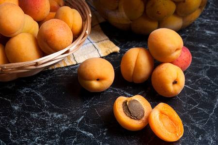 Ripe organic apricots harvested at summer time and big glass jar with canned sweet apricot compote on dark marble. Composition in rustic style - organic yellow juicy apricots whole and halved in yellow wooden basket and homemade apricot compote canned in big glass jar on dark marble background.の写真素材