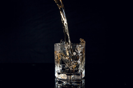 Whiskey pouring into glass with ice isolated on reflective black surface. Whiskey splash out of...