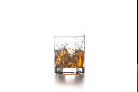 Big splash with drops of whisky from the glass of beverage with fallen ice cube into glass on light...