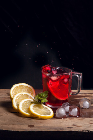 Summer iced drink - hibiscus cold tea with ice, lemon and mint.  Big splash with drops of hibiscus cold tea from the glass of beverage with fallen lemon slice into glass on wooden background. Hibiscus cold tea splash out of glass, many drops of beverage get out from glass.の写真素材