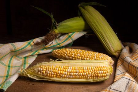 Two ears of ripe sweet corn with green leaves and green towel on vintage wooden background. Cobs with white and yellow grains.の写真素材