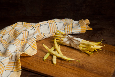 Cutting board with bunch and several pods of raw yellow pods of haricot and yellow kitchen towel on wooden background. French beans or Green beans also known as String beans from India. Vegetables collection.の写真素材