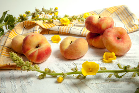 Several whole ripe chinese flat peach fruits or saturn peaches on vintage white wooden background with yellow kitchen towel and yellow flowers.の写真素材