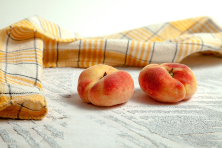 Two ripe chinese flat peach fruits or saturn peaches on vintage white wooden background with yellow kitchen towel.の写真素材