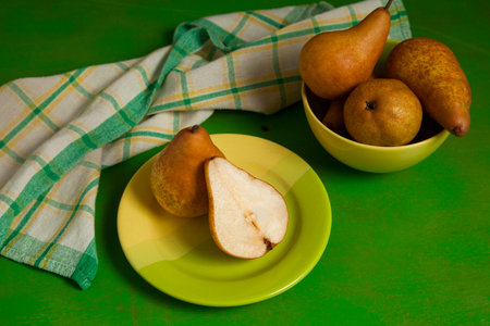 Green plate with whole and half of ripe juicy pear fruit and green kitchen towel on green wooden background.の写真素材