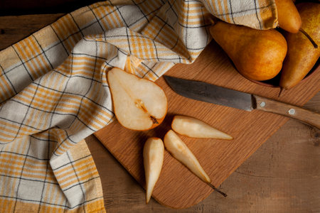 Wooden cutting board with slices and half of ripe juicy pear fruits, clay bowl with pears and yellow kitchen towel on vintage wooden background.の写真素材