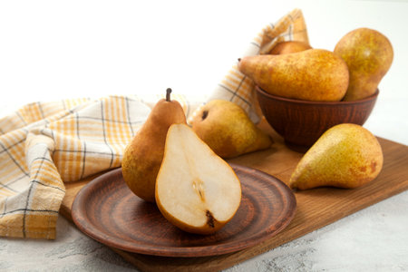 Clay plate with whole and half of ripe juicy pear fruits, wicker basket with pears and yellow kitchen towel on wooden cutting board.の写真素材