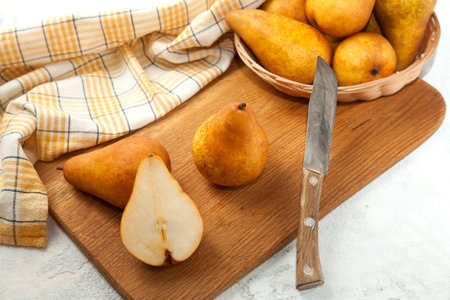 Wooden cutting board with whole and halves of ripe juicy pear fruits, wicker basket with pears and yellow kitchen towel on white wooden background.の写真素材