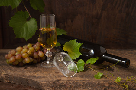 The concept of a delicious alcoholic drink, wine and grapes. Wine bottle, empty glass and glass of white wine and bunch of pink grapes on dark rustic wooden background with grapes leaves.の写真素材